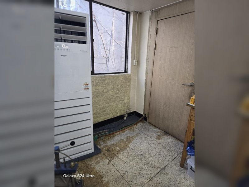 오래된 스탠드형 에어컨 폐기물 수거 작업이 완료되어 공간이 정리되었지만, 바닥에 약간의 이물질이 남아있다.<br>